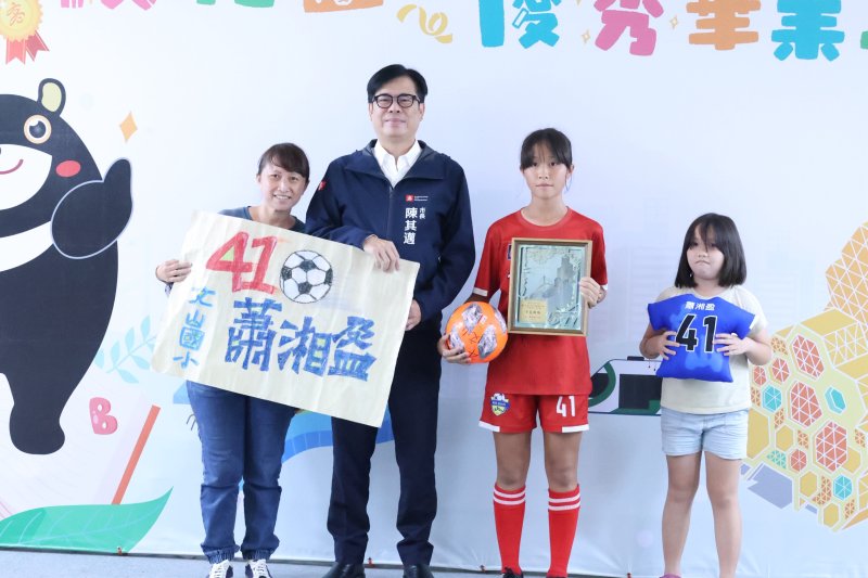 文山國小畢業生蕭湘盈(右二)穿著學校足球隊服與市長合影,她曾與隊友勇奪全國女子足球賽季軍,母親也驕傲地手持印有女兒姓名的應援海報,共同見證這份榮耀。 文山國小畢業生蕭湘盈(右二)穿著學校足球隊服與市長合影,她曾與隊友勇奪全國女子足球賽季軍,母親也驕傲地手持印有女兒姓名的應援海報,共同見證這份榮耀。